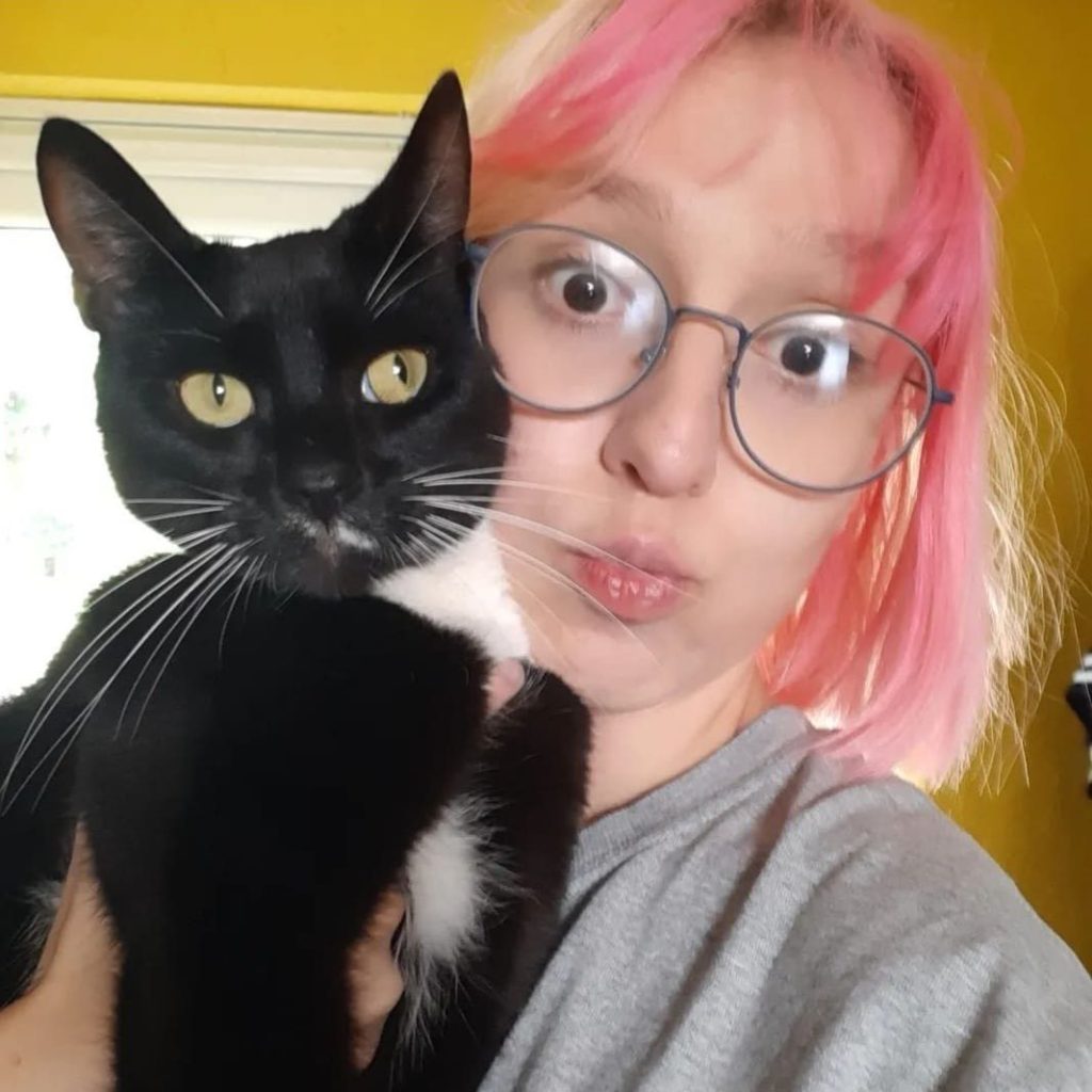Rhi Armstrong, a white woman with pink hair, poses with a black cat on her shoulder.